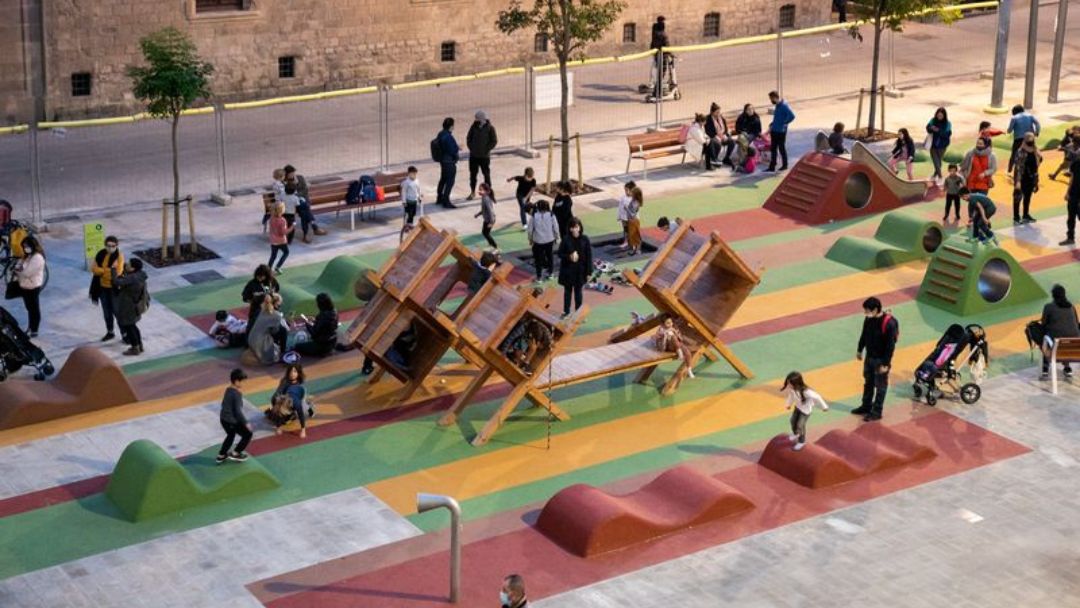 efecto-ikea-urbano-participacion-ciudadana-plaza-juegos Vista aérea de una plaza pública colorida con zonas de juego de madera y personas disfrutando, ejemplo de los beneficios del Efecto IKEA urbano y la participación ciudadana.