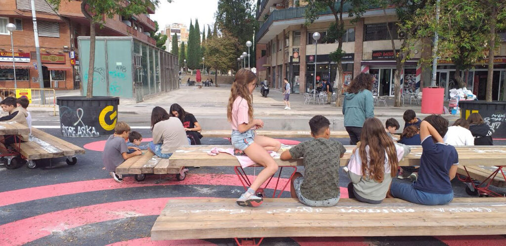 Niños y jóvenes utilizan un espacio público transformado con bancos y mesas en la calle.