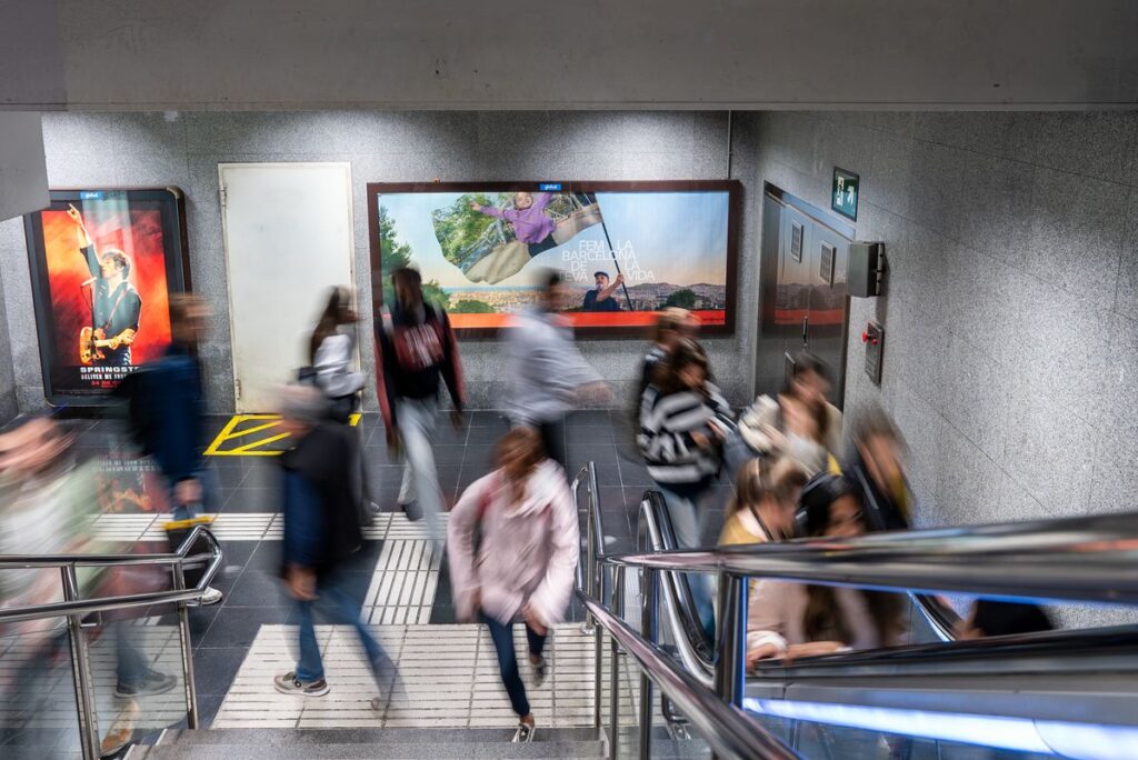 Pasajeros descendiendo en la estación de metro de Barcelona ante un soporte publicitario institucional sobre calidad de vida urbana. Commuters walking through a Barcelona subway station past a municipal billboard highlighting urban quality of life.